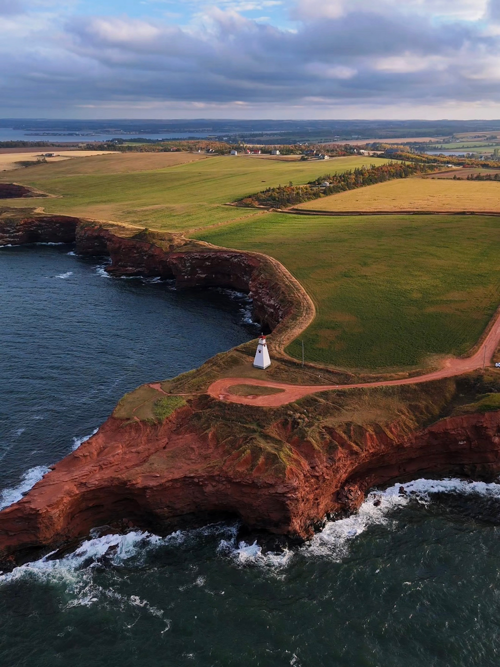 Scenic aerial view of Cape Tryon Lighthouse on Prince Edward Island &mdash; dramatic red cliffs and ocean waves at sunset.