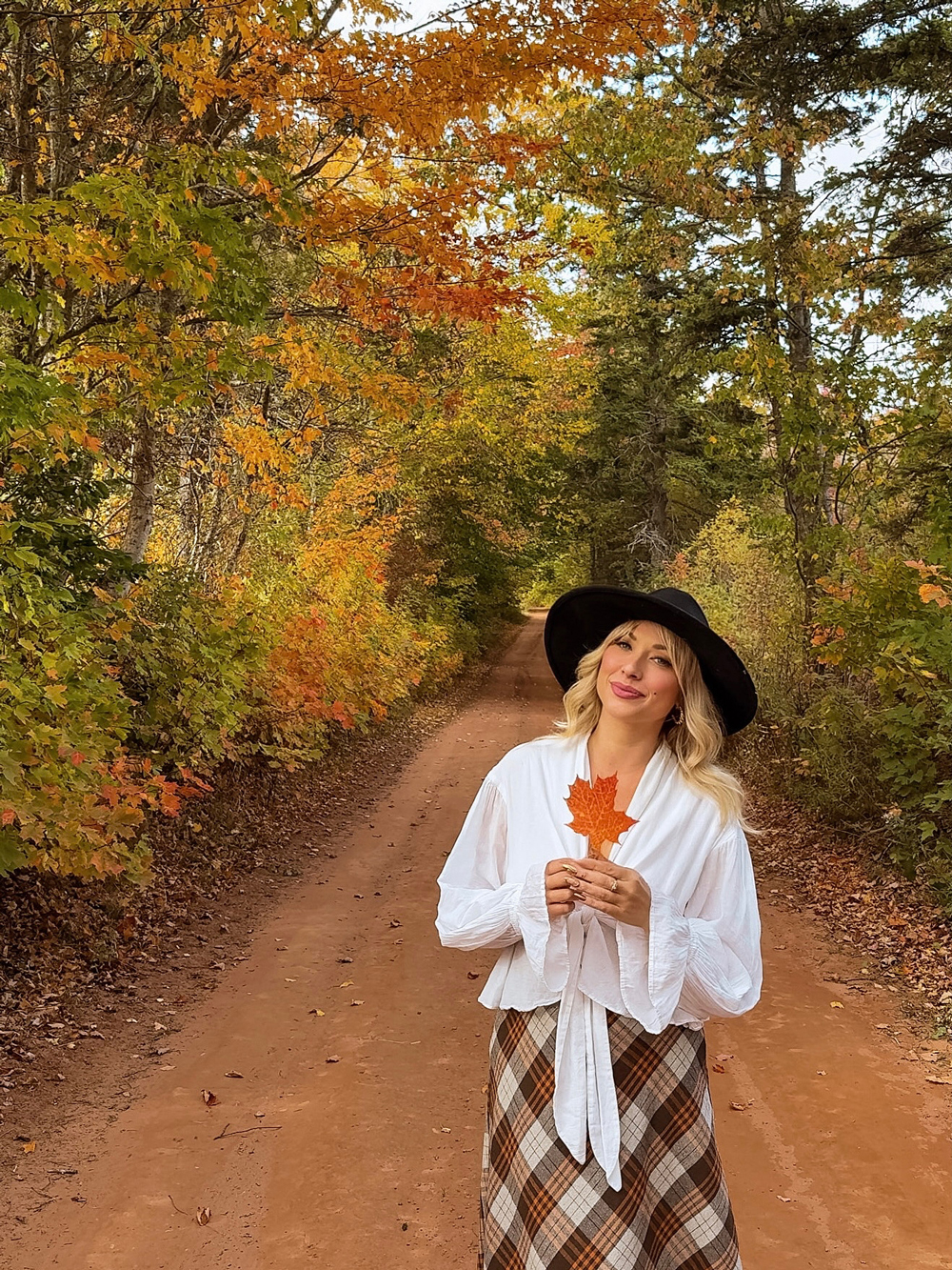 Fall colors along a red dirt road near Charlottetown, PEI &mdash; woman holding a maple leaf surrounded by golden foliage.