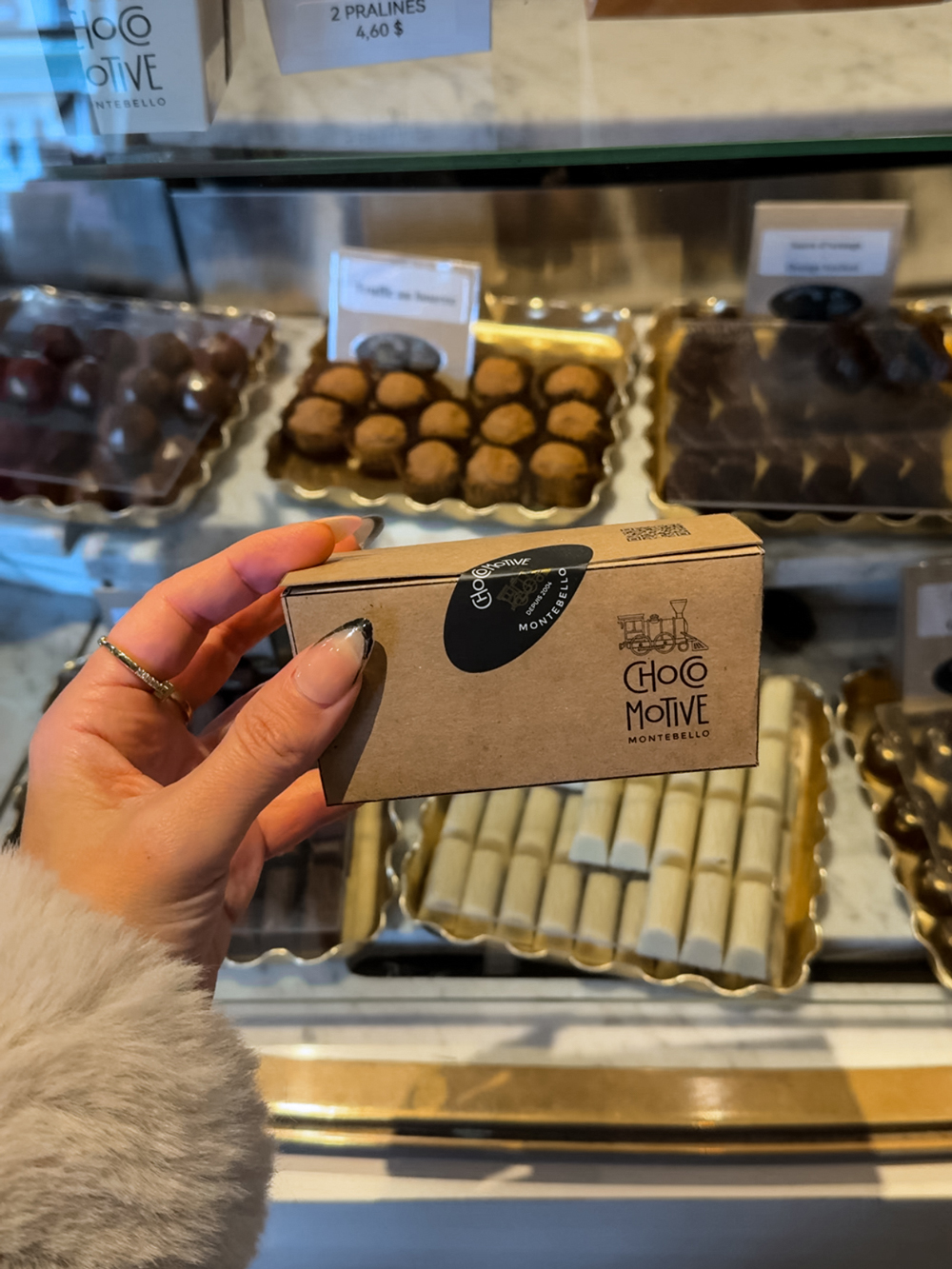 Hand holding a small box of chocolates at ChocoMotive Montebello, with trays of truffles and handmade holiday sweets in the background.