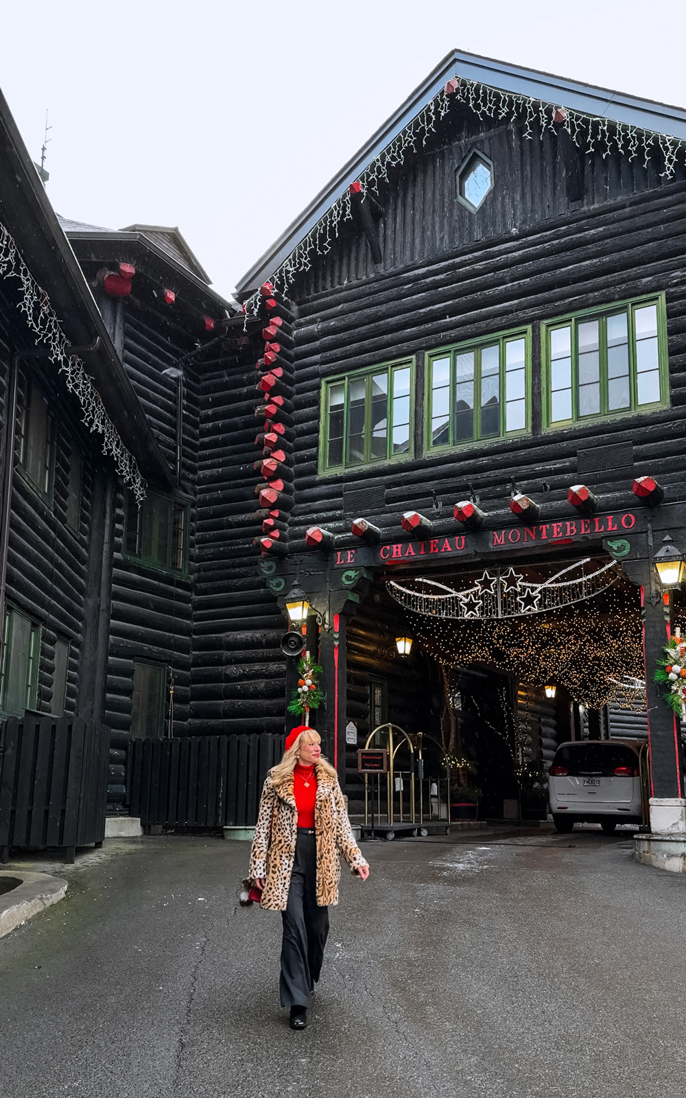 Entrance of Fairmont Le Ch&acirc;teau Montebello decorated with Christmas lights and garlands as a woman walks outside wearing a leopard coat and red beret.