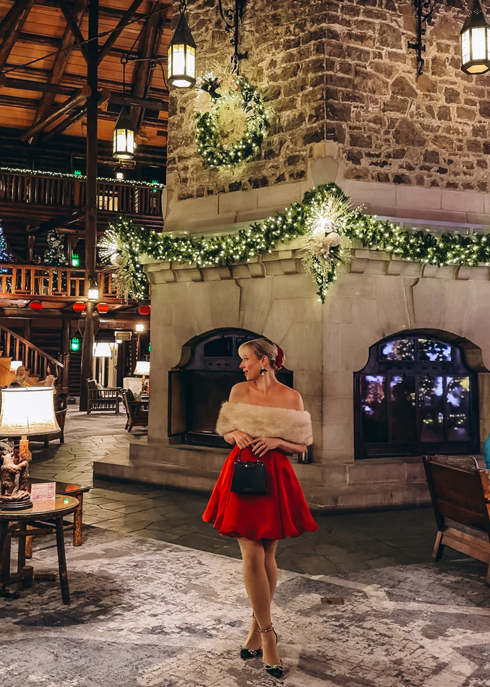 Woman in a red holiday dress standing beside the large decorated stone fireplace at Fairmont Le Ch&acirc;teau Montebello during Christmas.