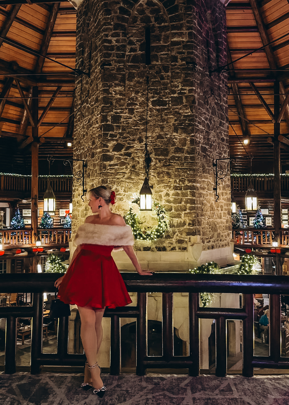 Woman in a red festive dress standing beside the stone fireplace on the mezzanine inside Fairmont Montebello, surrounded by garlands and holiday lights.
