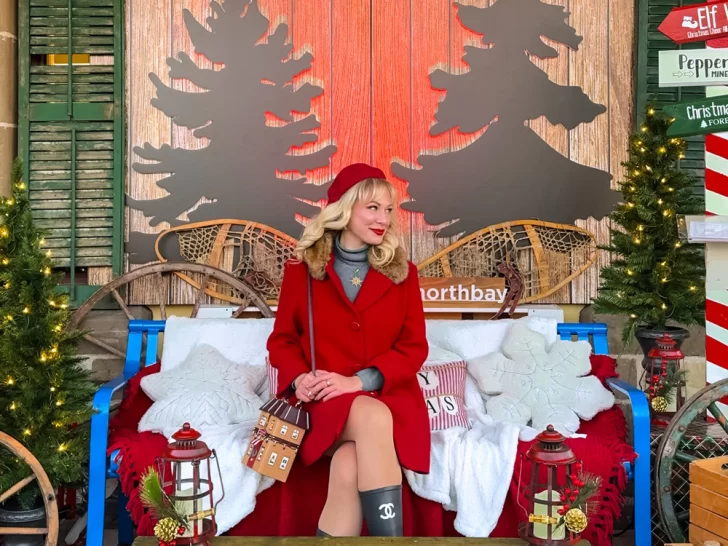 Woman in a red coat sitting on a decorated bench with snowshoes, lanterns, and Christmas trees at the North Bay Winter Market, one of the top things to do in North Bay at Christmas time.
