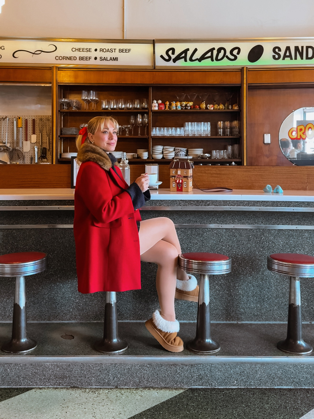 Woman in a red coat sitting at a retro diner counter in Crossroads Convenience, with red stools and a Christmas toy shop shaped purse beside her.
