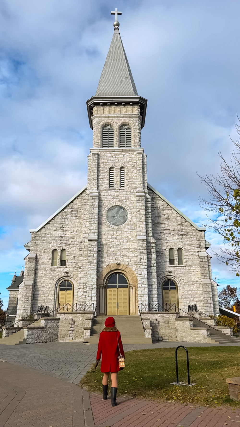Woman in a red coat walking toward a large stone church in North Bay, with a tall steeple and cross.