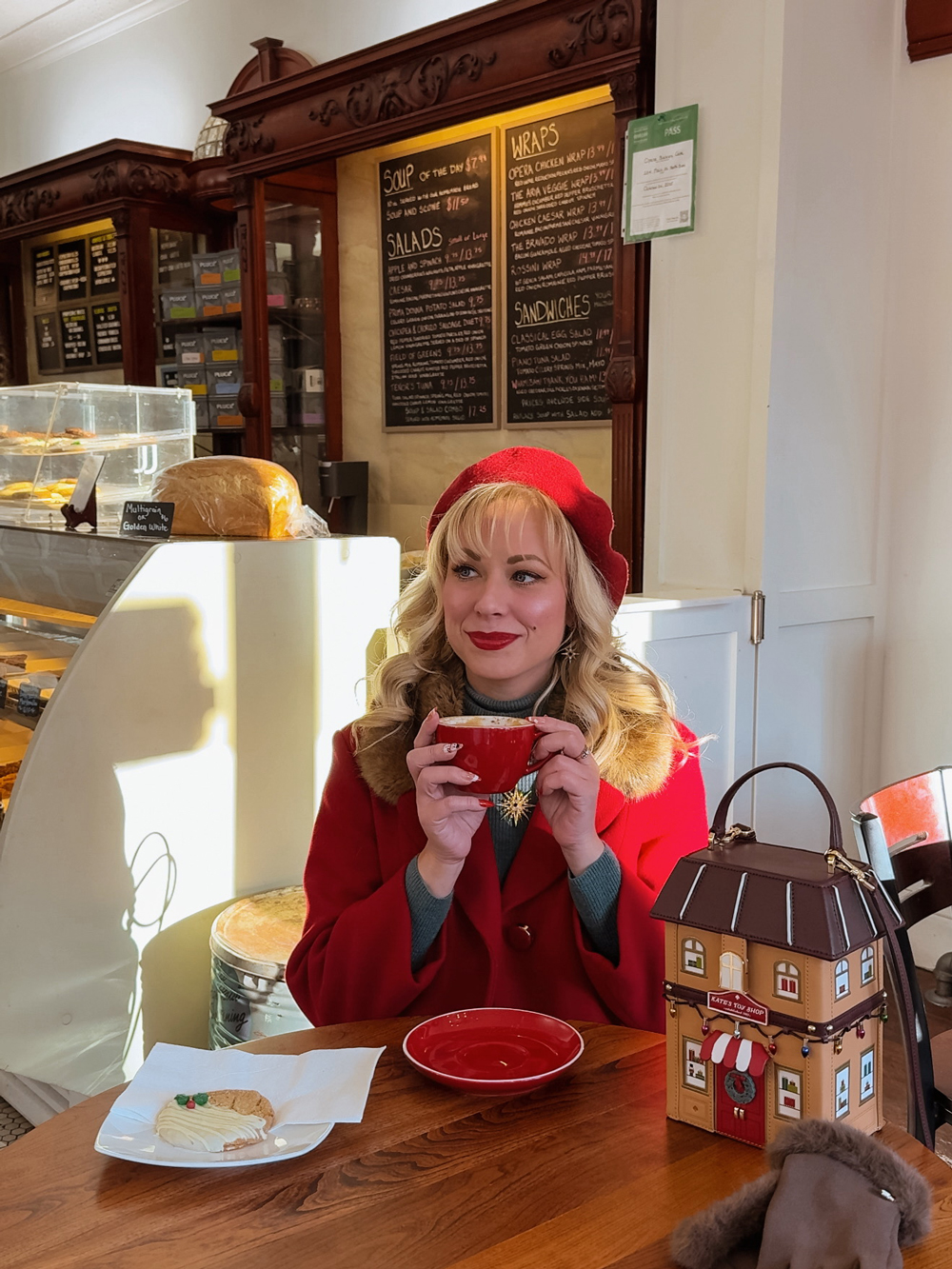 Woman in a red coat holding a red mug inside Opera Bakery Caf&eacute; with a cookie and house-shaped purse on the table.