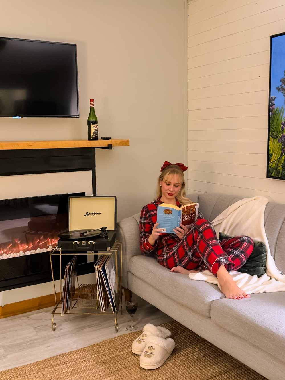 Woman in plaid pajamas reading on a grey sofa beside an electric fireplace and a record player at The Finch Beach Resort in North Bay.