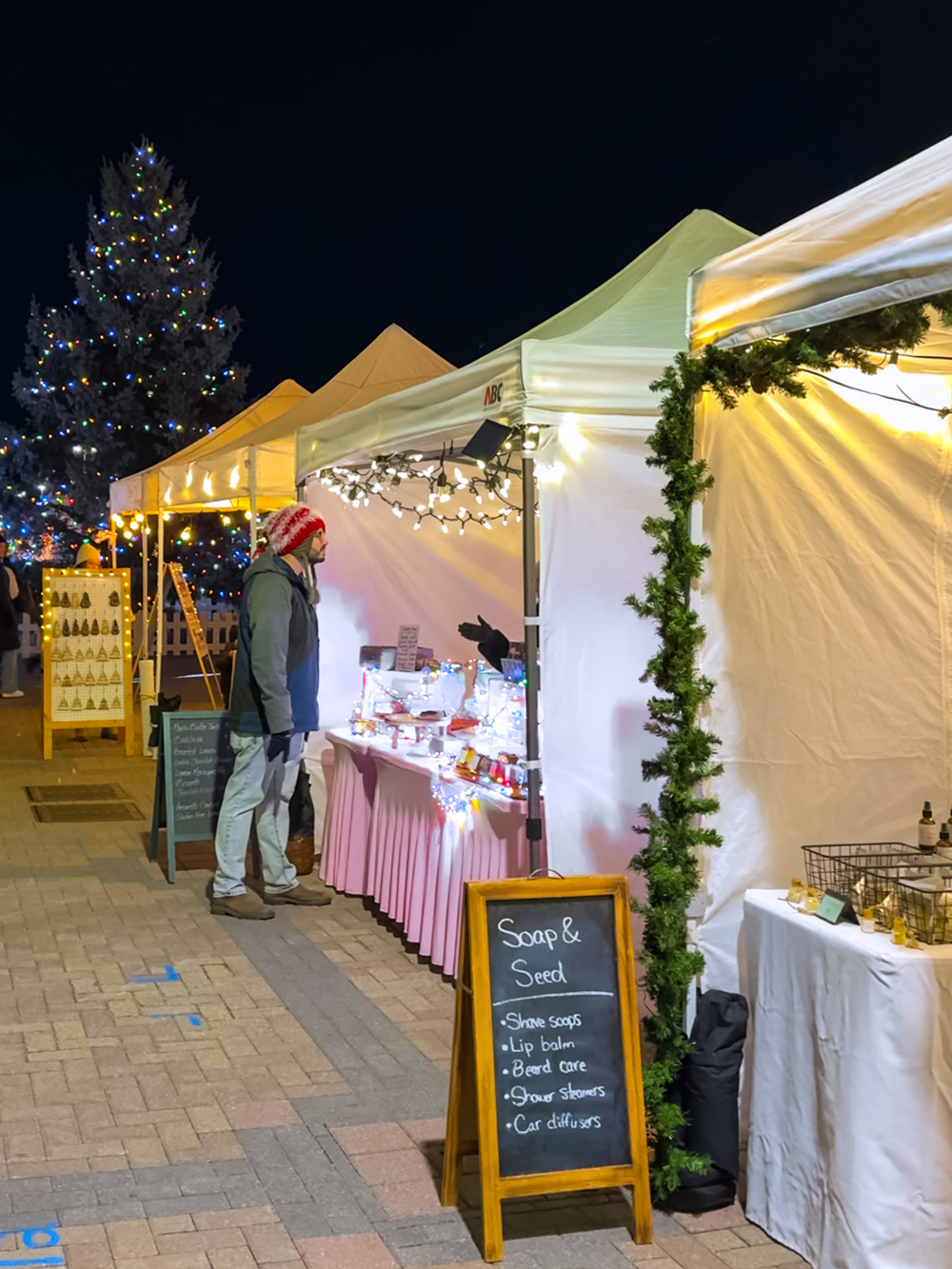 Holiday market tents lit with string lights, with shoppers browsing and a decorated Christmas tree in the background at the North Bay Winter Market.
