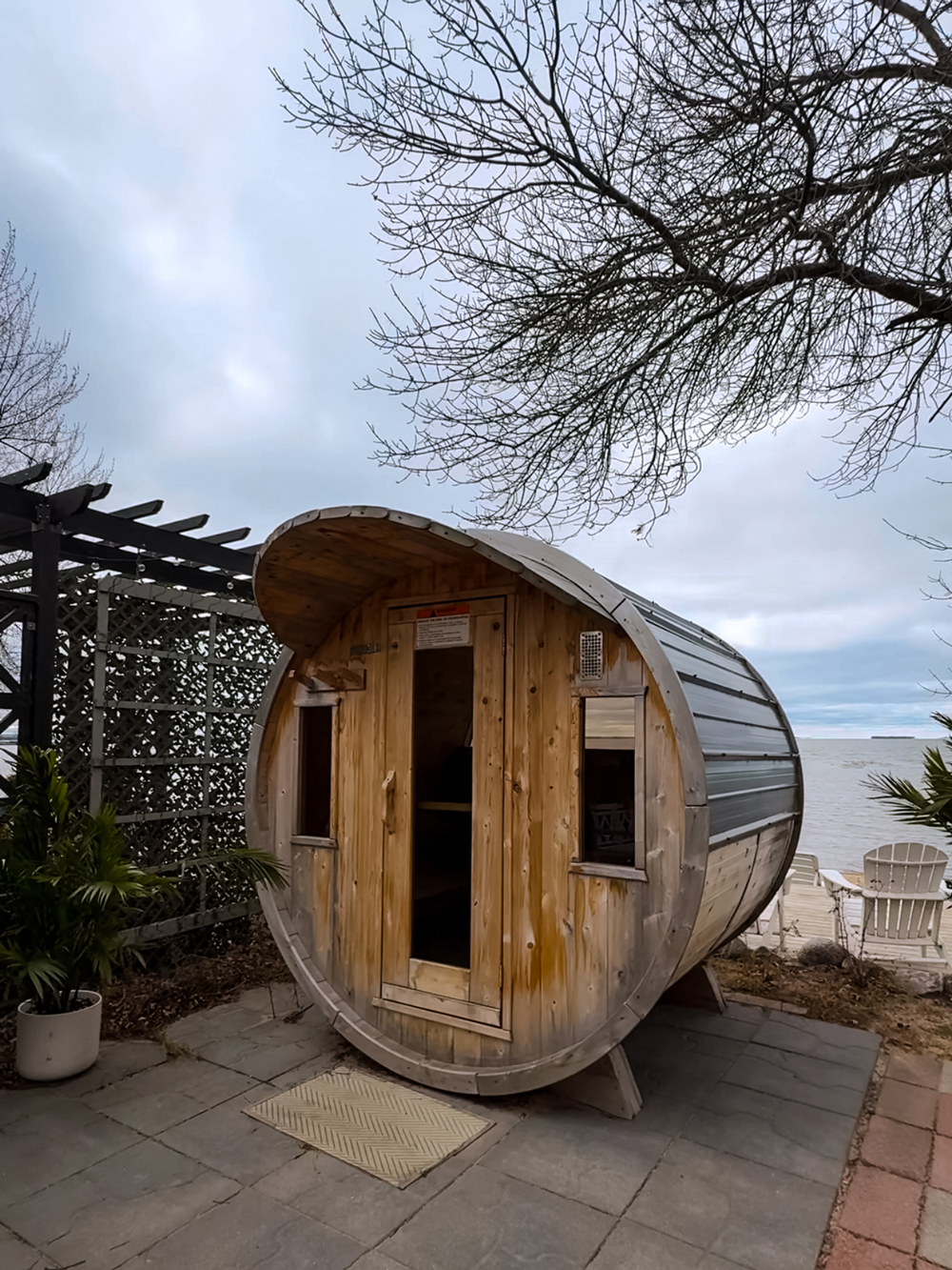 Wooden barrel sauna at The Finch in North Bay, set on a lakeside patio with bare trees and Adirondack chairs nearby.