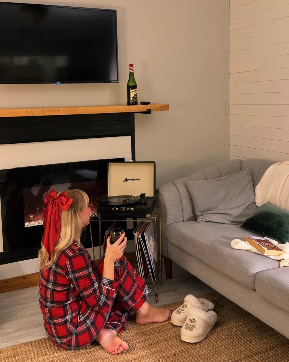 Woman in plaid pajamas sitting on the floor at The Finch Resort in North Bay with a glass of wine, beside a record player and fireplace.