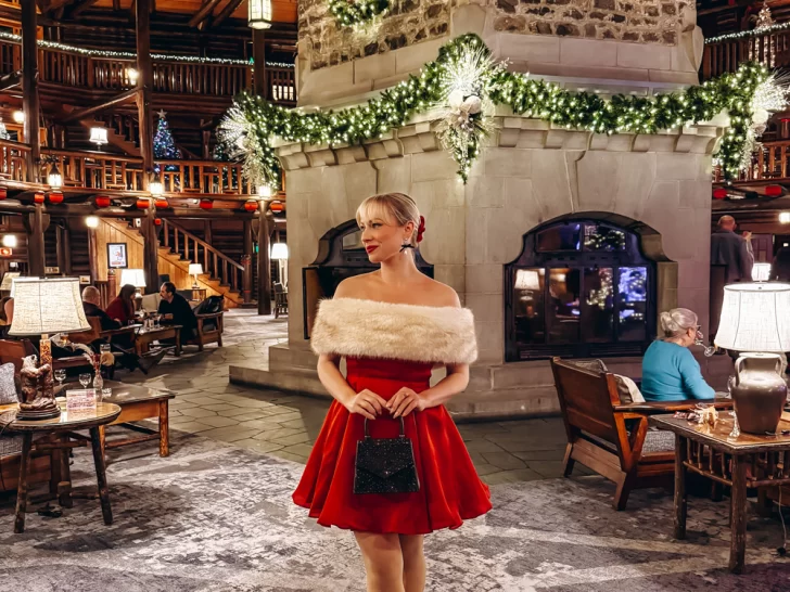 Woman in a festive red holiday dress standing in front of the decorated stone fireplace at Fairmont Le Ch&acirc;teau Montebello during Christmas. One of the most magical things to do in Montebello at Christmas.