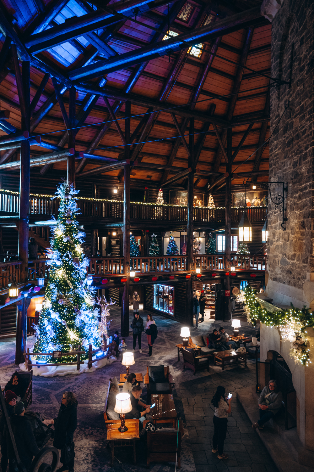 Grand Christmas tree and festive decorations inside the lobby of Fairmont Le Ch&acirc;teau Montebello, with guests relaxing beneath the log-cabin architecture.