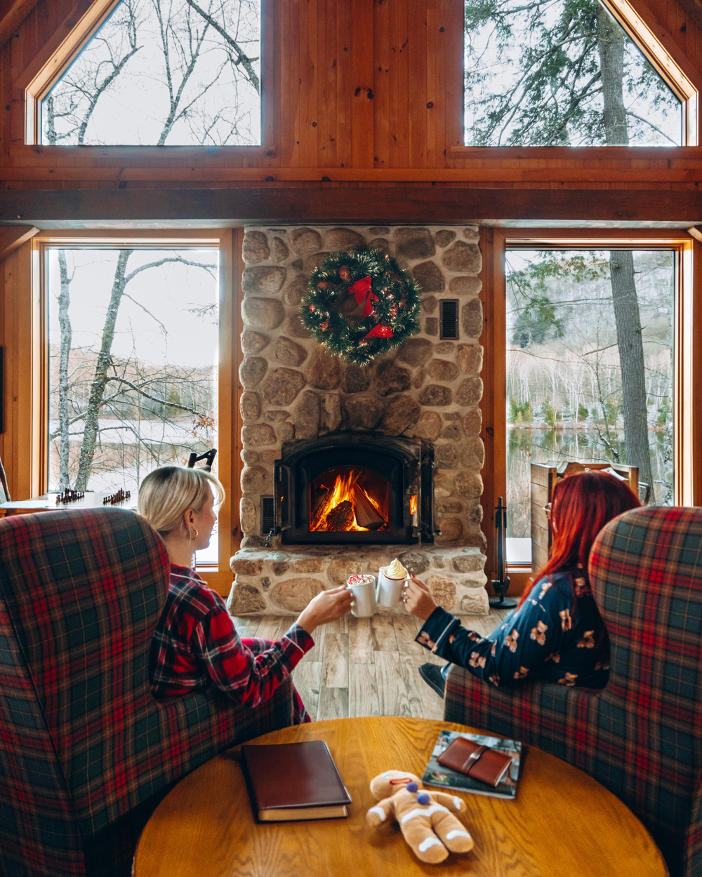 Two women in plaid pajamas clinking mugs of hot chocolate in front of a stone fireplace inside a Kenauk Nature chalet decorated for Christmas.