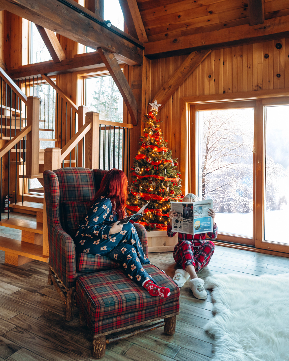 Two women relaxing in Christmas pajamas beside a decorated tree inside a Kenauk Nature chalet.