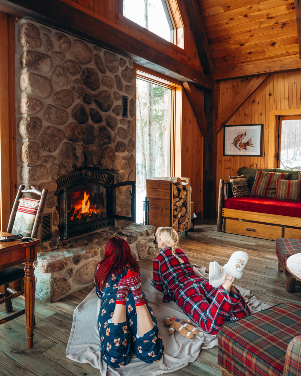 Two women lying on a blanket in front of the stone fireplace inside a Kenauk Nature chalet, enjoying a cozy winter morning.