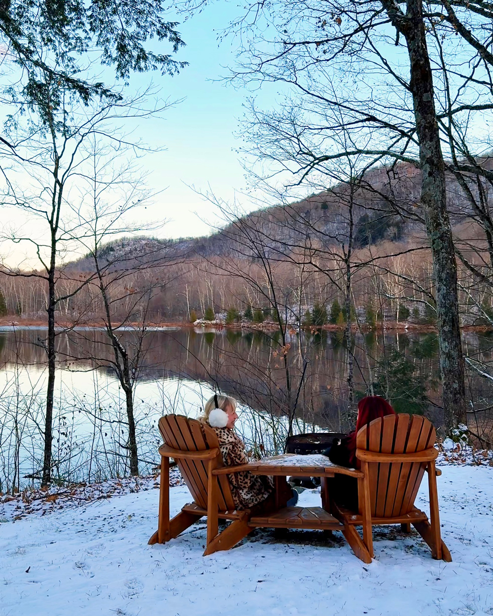 Two women sitting in Adirondack chairs beside a snowy lakeside at Kenauk Nature, surrounded by peaceful forest views.