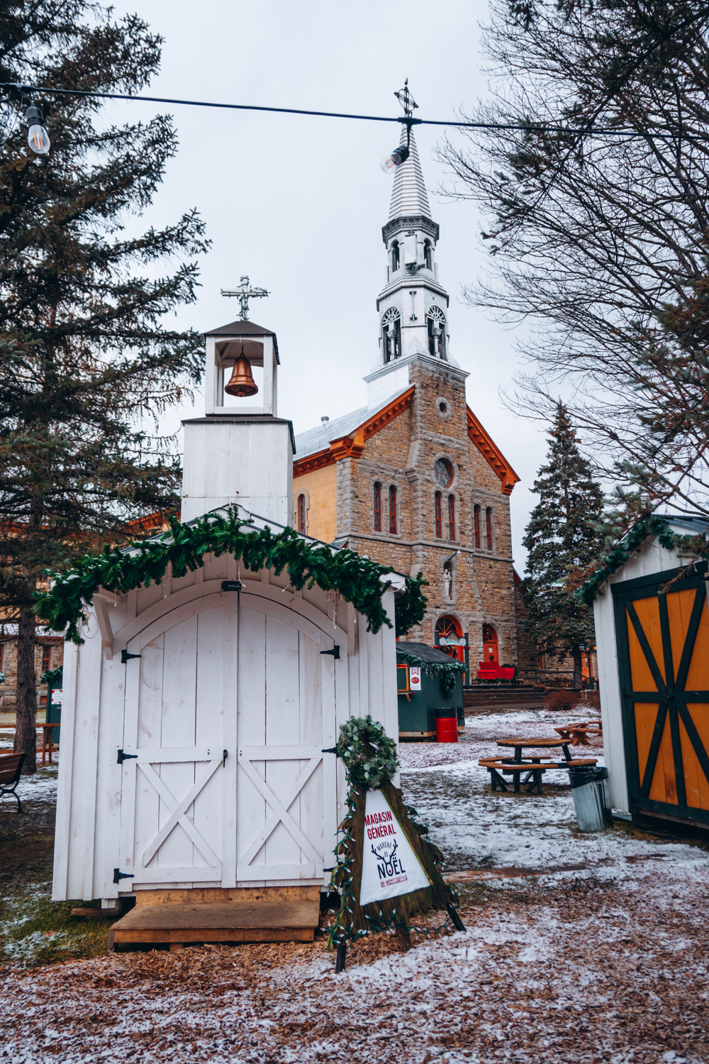 A small white wooden chalet-style booth decorated with greenery at the Montebello Christmas Market, with the village church in the background.