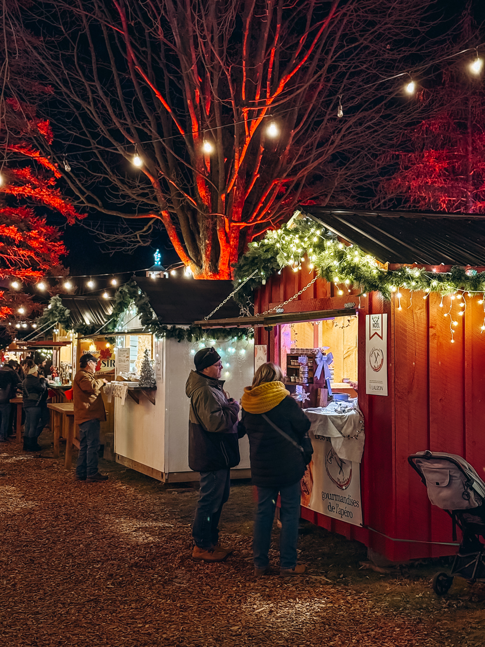 People browsing festive vendor stalls decorated with lights and garlands at the Montebello Christmas Market at night.