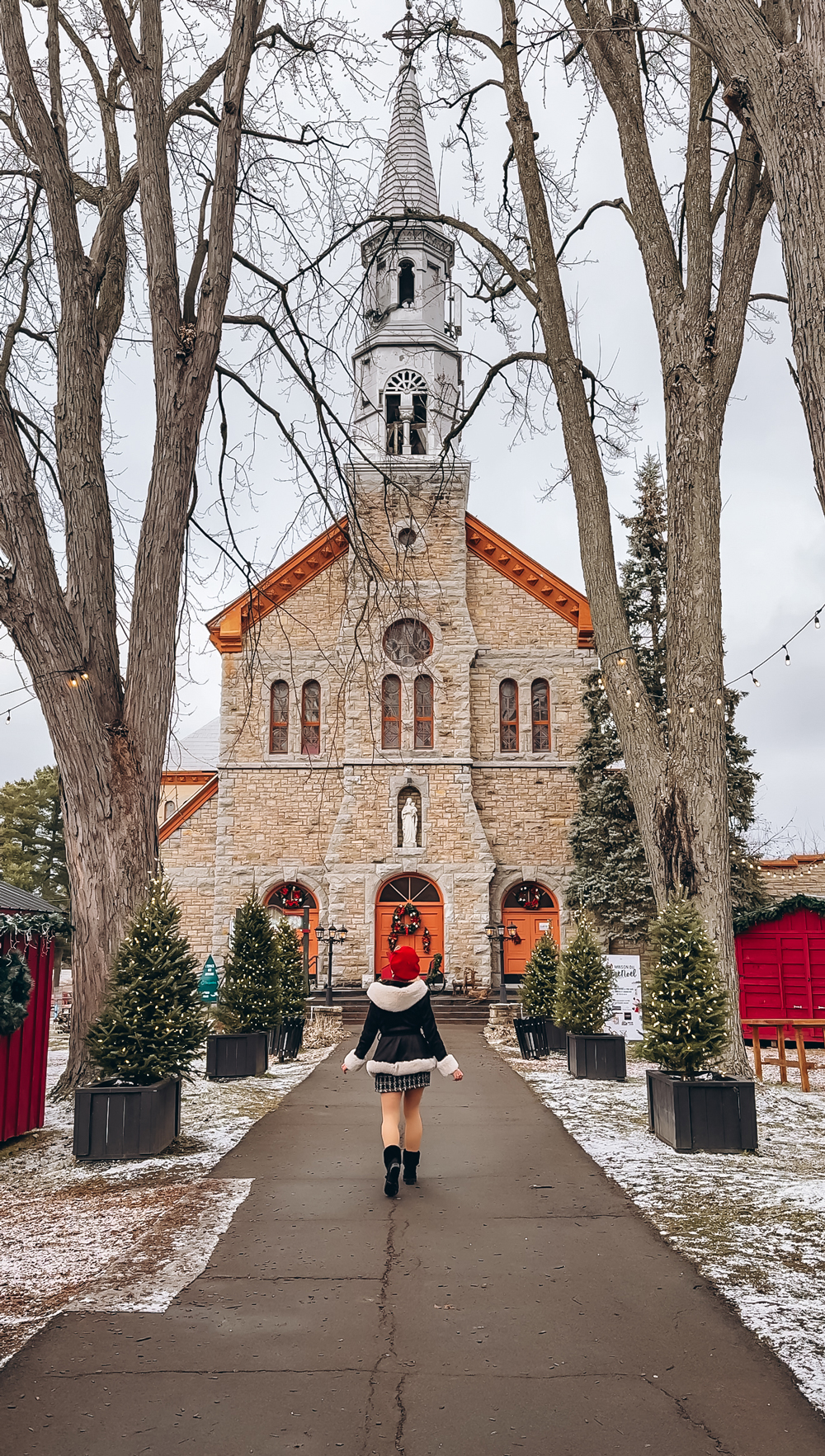 A woman in festive winter clothing walking toward Montebello&rsquo;s historic stone church decorated for Christmas.